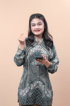 Asian woman wearing elegant batik outfit holding smartphone and showing finger heart gesture, smiling with confident and friendly expression, symbolizing culture, positivity, and modern lifestyle.の写真素材