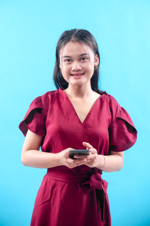 A young Asian woman in a red dress smiling while holding a smartphone against a blue background,...