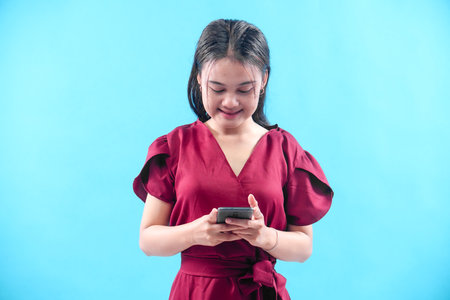 A young Asian woman in a red dress smiling while looking down at her smartphone, representing...