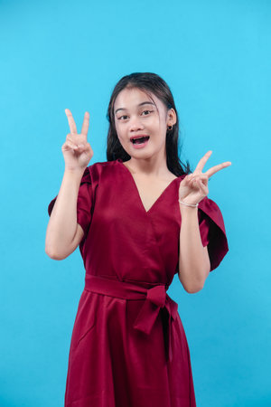 A cheerful young woman wearing a red dress shows a double peace sign with both hands, smiling brightly with playful expression, standing against a plain blue background.の写真素材