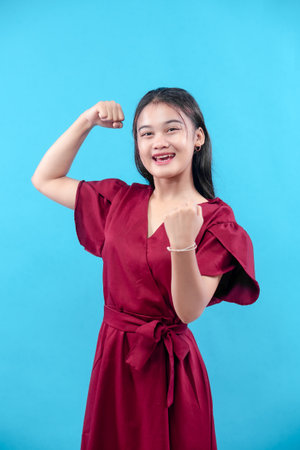 A confident young woman in a red dress poses with a powerful gesture, smiling brightly and showing strength, optimism, and motivation against a blue background.の写真素材