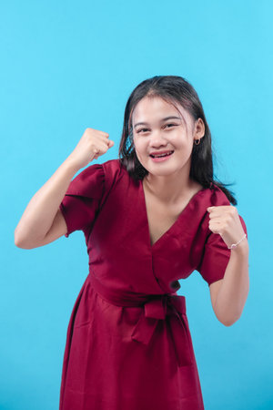 A cheerful young woman in a red dress poses with raised fists and a bright smile, expressing confidence, positivity, and determination against a vibrant blue background.の写真素材