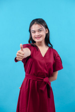 Smiling young woman in red dress showing thumbs up gesture, isolated on blue background, representing approval, success, positivity, confidence, motivation, agreement, and encouragement.の写真素材