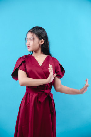 A young woman in a red dress raising both hands with a serious face, isolated on blue background, showing rejection gesture and strong attitude.の写真素材