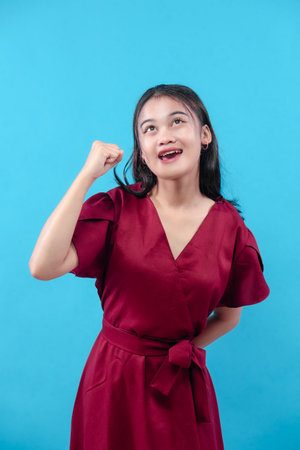 A cheerful young woman in a red dress raises her fist with a joyful smile, expressing confidence, success, and motivation against a clean blue background.の写真素材