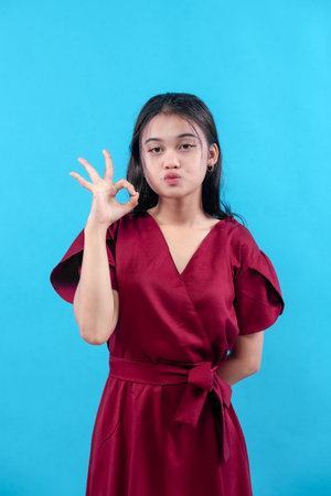 A young woman wearing a stylish red dress poses with a confident expression, making a hand gesture, standing against a bright blue background for creative concepts.の写真素材