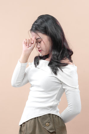 An Asian woman in a white blouse stands with her eyes closed and hand on her forehead, expressing stress and deep thought, symbolizing headache, fatigue, or emotional pressure.の写真素材