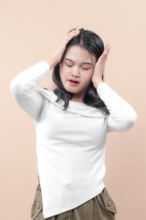An Asian woman in a white blouse touching her head with both hands, eyes closed and lips slightly open, expressing stress, headache, or mental exhaustion.の写真素材
