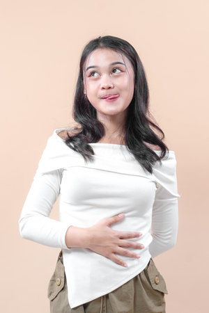 A young Asian woman in a white top touches her stomach while smiling playfully, looking up with a thoughtful expression that conveys curiosity and positivity.の写真素材