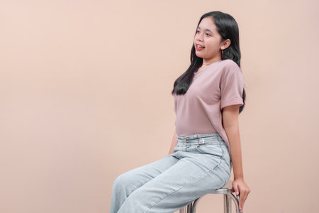 Young Asian woman with long black hair wearing pastel t-shirt and denim jeans, sitting casually on chair, smiling with natural expression, showing confidence and modern casual style.の写真素材
