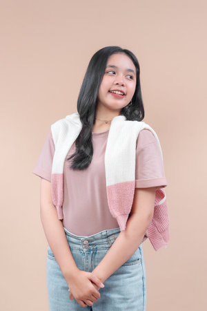 Asian woman with long black hair wearing pastel pink t-shirt, denim jeans, and knit sweater on shoulders, smiling softly while looking to the side with casual modern charm.の写真素材