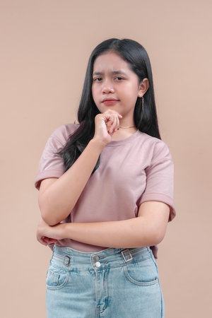 An Asian woman in a casual pink t-shirt and denim jeans stands with a thoughtful pose, hand on chin,...