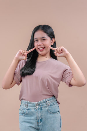 An Asian woman in a casual t-shirt and jeans smiling confidently while pointing to her cheeks with...