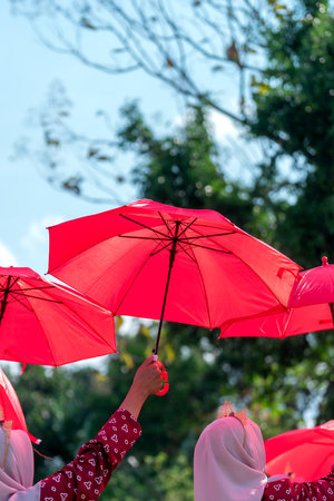 A woman in a white hijab raises a bright red umbrella under the blue sky, creating a vivid outdoor...