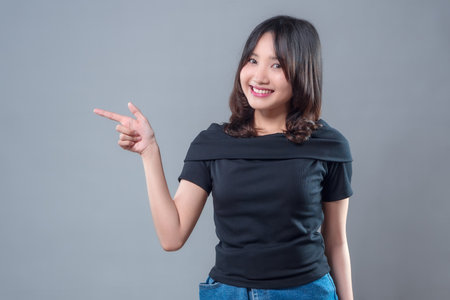 A cheerful Asian woman smiling confidently while pointing to the side with her finger, wearing a black top and blue jeans, perfect for business presentation, advertising, or promotional concepts.の写真素材