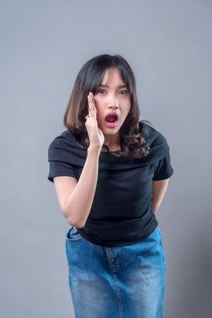 An Asian woman looks surprised and shouts with her mouth open, expressing shock or calling out. She wears casual clothes, showing emotion, communication, and expressive human reaction.の写真素材