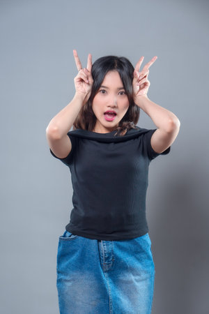 A cheerful Asian woman with long hair making a playful gesture with her fingers shaped like bunny ears. She wears a black shirt and denim skirt, expressing surprise, fun, and youthful energy.の写真素材