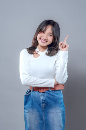 A cheerful young Asian woman smiling brightly while pointing upward with one finger, wearing a white long-sleeve top and denim skirt, expressing confidence and positive idea inspiration.の写真素材