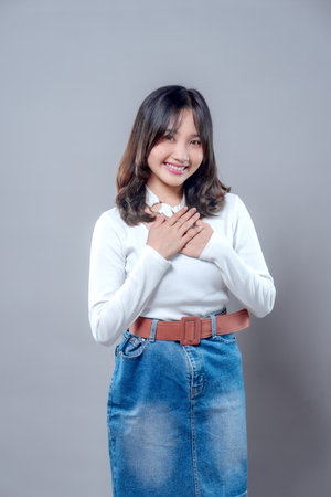 A beautiful young Asian woman smiling gently with her hands on her chest, wearing a white sweater and denim skirt, expressing gratitude, kindness, and confidence in a calm and friendly manner.の写真素材