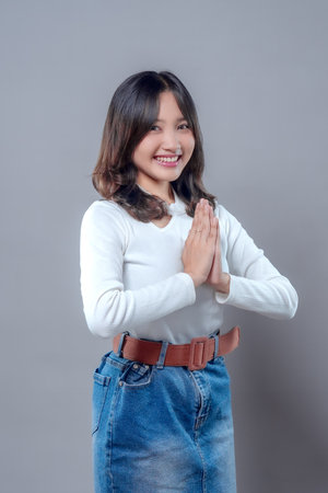 A beautiful Asian woman smiling warmly while pressing her palms together in a greeting gesture, wearing a white long sleeve top and denim skirt, showing kindness, respect, and positive emotion.の写真素材