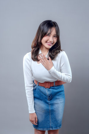 A cheerful young Asian woman smiling warmly while placing her hand on her chest, expressing gratitude, politeness, and sincerity. She wears a white top and denim skirt in a friendly and respectful pose.の写真素材