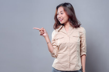 A smiling Asian woman looking sideways while pointing with her finger. She wears a beige shirt and gray pants, showing a confident, friendly, and approachable attitude suitable for business and advertising.の写真素材