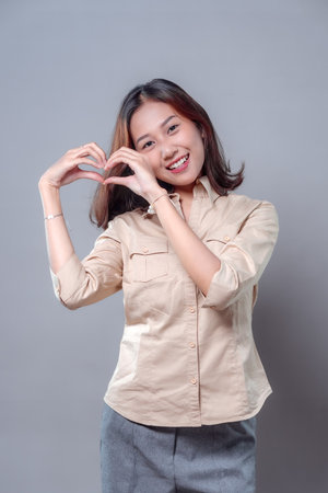 A smiling Asian woman forming a heart shape with her hands, expressing kindness, positivity, and affection. She wears a beige shirt and gray pants, radiating warmth, charm, and confidence in a friendly pose.の写真素材