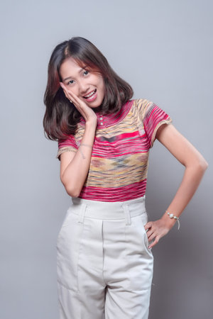 A cheerful Asian woman smiling with a surprised expression while touching her face. She wears a...