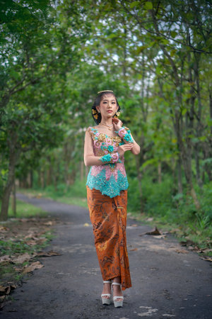 An elegant Indonesian woman wearing a colorful kebaya and batik skirt walks gracefully on a rural...