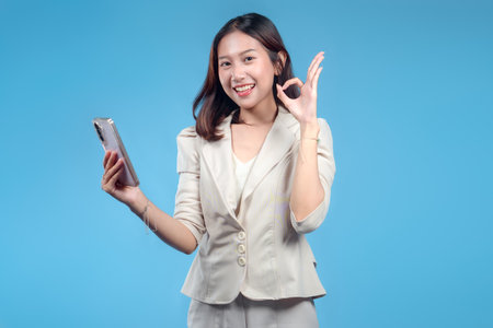 An Indonesian businesswoman smiling confidently while holding a smartphone and making an OK gesture. Her formal attire and positive expression portray a strong, professional image suitable for global corporate and digital business content.の写真素材
