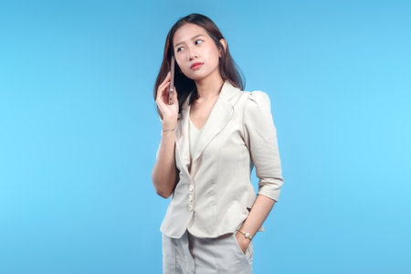 A thoughtful Indonesian businesswoman holding a smartphone while looking away, dressed in professional attire on a clean blue background, showing a focused corporate concept.の写真素材