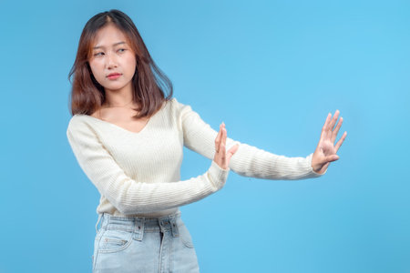 An Indonesian woman with a cautious expression extending her hands sideways in a defensive gesture, wearing a cream sweater and jeans against a clean blue background.の写真素材
