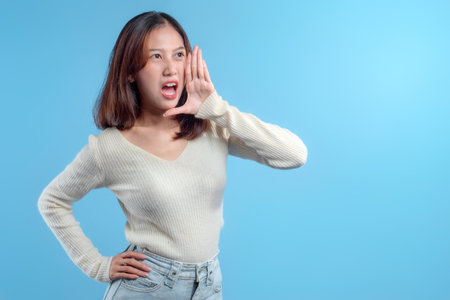 A confident Indonesian woman making a shouting gesture with one hand near her mouth, expressing energy and communication in a casual outfit against a clean blue background.の写真素材