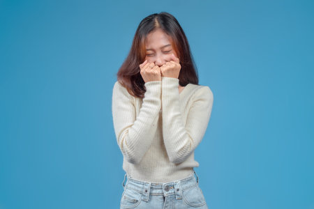 A woman with a shy, adorable expression, eyes closed and hands near her face, wearing a cream sweater and jeans against a clean blue background, conveying charm and warmth.の写真素材