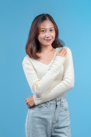A woman smiling confidently while touching her shoulder, wearing a cream sweater and jeans against a blue background, showing a calm and friendly demeanor.の写真素材