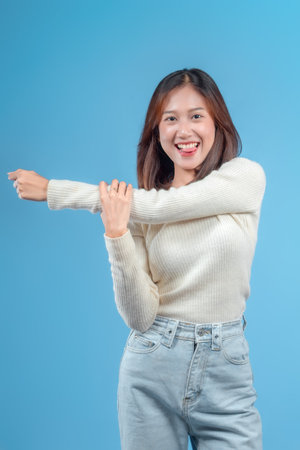 An Indonesian woman stretching her arm with a cheerful, playful expression, wearing a cream sweater and jeans against a clean blue background, conveying energy and positivity.の写真素材