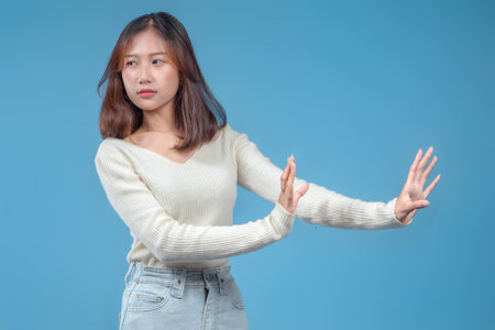 An Indonesian woman with a cautious expression extending her hands outward in a defensive gesture, wearing a cream sweater and jeans against a clean blue background.の写真素材