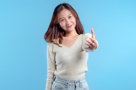 A friendly Indonesian woman reaching her hand forward with a warm smile, expressing welcome, connection, and positive interaction against a bright blue background.の写真素材