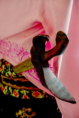 Close up detail of traditional Javanese dagger or Keris tucked in the back of groom attire featuring pink beskap and batik sash.の写真素材