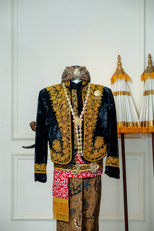 Traditional Javanese groom wedding attire featuring black velvet beskap with gold embroidery, necklace, and blangkon displayed on mannequin.の写真素材