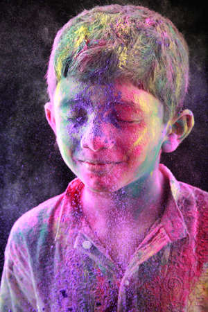 A boy plays Holi with colored powder exploding around his face in a dark background.