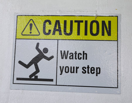 Yellow and black warning sign on stairs stating "Watch your Step" signifying a potential trip hazard, steep stairs and care to be taken when going up or down stairsの写真素材