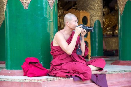 Buddhist monk in traditional robes is taking photos by camera at the Shwedagon Pagoda, a gilded stupa located in Yangon, Myanmar. The 99 metres tall pagoda is situated on Singuttare Hill, to the West of Kandawgyi Lake and dominates the Yangon skyline. It のeditorial素材