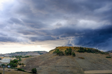 A view of a hill with scattered trees, framed by dramatic, stormy clouds over a rural landscape at sunset.の写真素材
