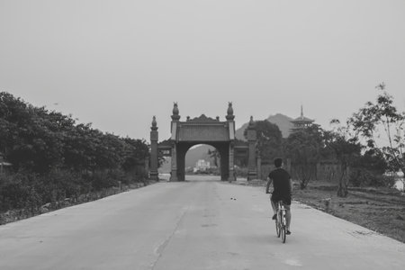 A lone cyclist rides towards a traditional Vietnamese gate in a serene, empty road scene. The black-and-white tones emphasize the peaceful ambiance.の写真素材