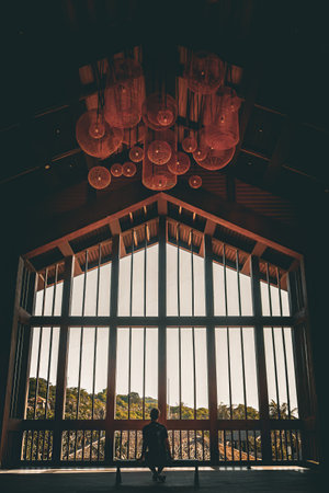 Silhouetted figure seated by a large window with intricate hanging lanterns above, gazing out at a scenic landscape.の写真素材