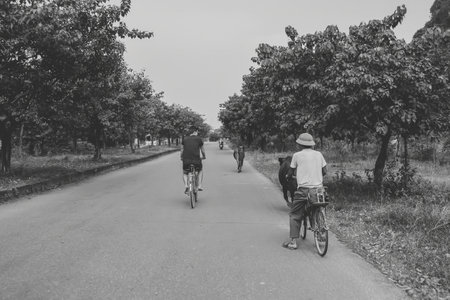A black-and-white photograph captures two men riding bicycles down a quiet rural road lined with...