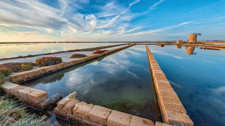 Salt pans near Trapani, Sicily, with still water reflecting the sky and an old windmill ruin at sunset.の写真素材