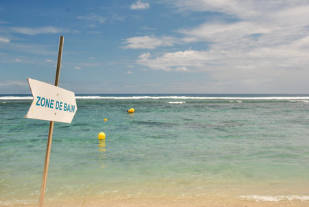 Sandy beach with a "Zone de Bain" sign in French pointing towards a calm turquoise sea with floating yellow buoys under a partly cloudy sky.の写真素材