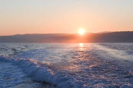 Calm sea at sunset with soft waves and distant hills, glowing in warm tones of orange and purple under a clear sky.の写真素材